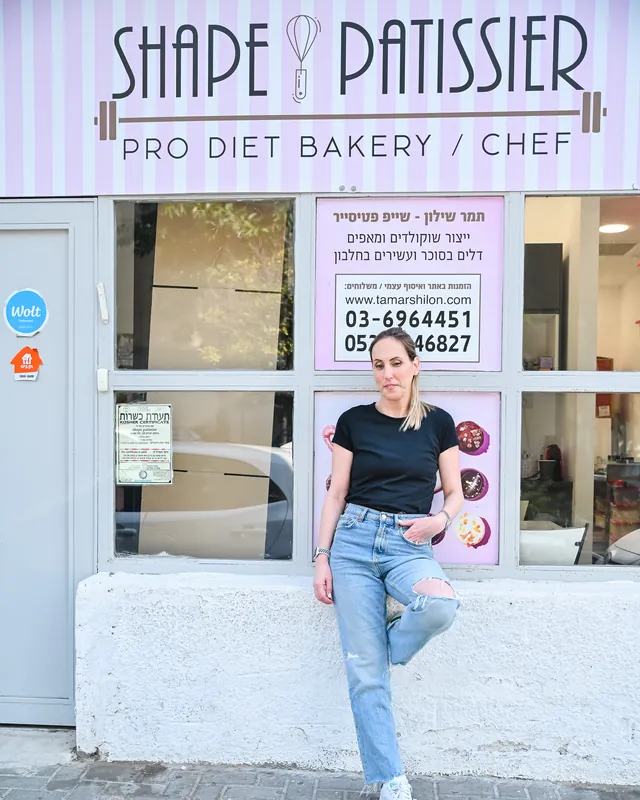 תמר שילון עומדת מול חזית המפעל של Shape Patissier בתל אביב, עם השלט מעל הכניסה